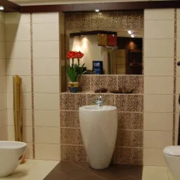 Jasna łazienka z beżowymi i brązowymi płytkami, wolnostojącą umywalką, bidetem, toaletą i drewnianą figurką żyrafy jako elementem dekoracyjnym.